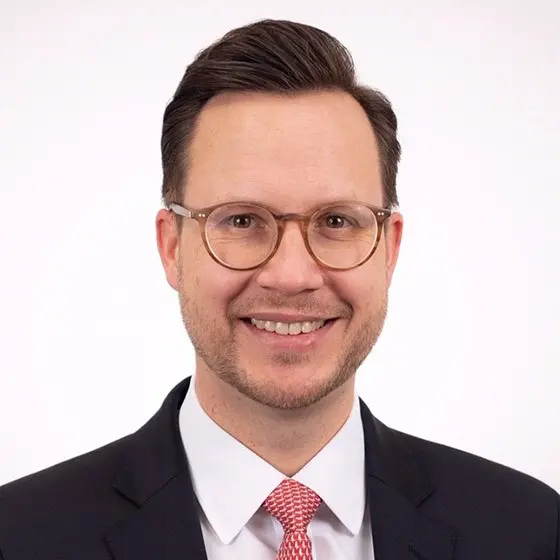 Portrait von Christian Stockfleth. Er trägt einen, Anzug, weißem Hemd und roter Krawatte, lächelt vor hellem Hintergrund, blickt direkt in die Kamera.
