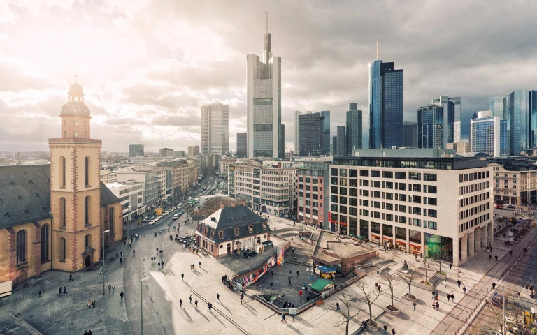 Stadtansicht mit Kirche, Platz mit Personen und moderner Skyline im Hintergrund bei Tageslicht und teils bewölktem Himmel.